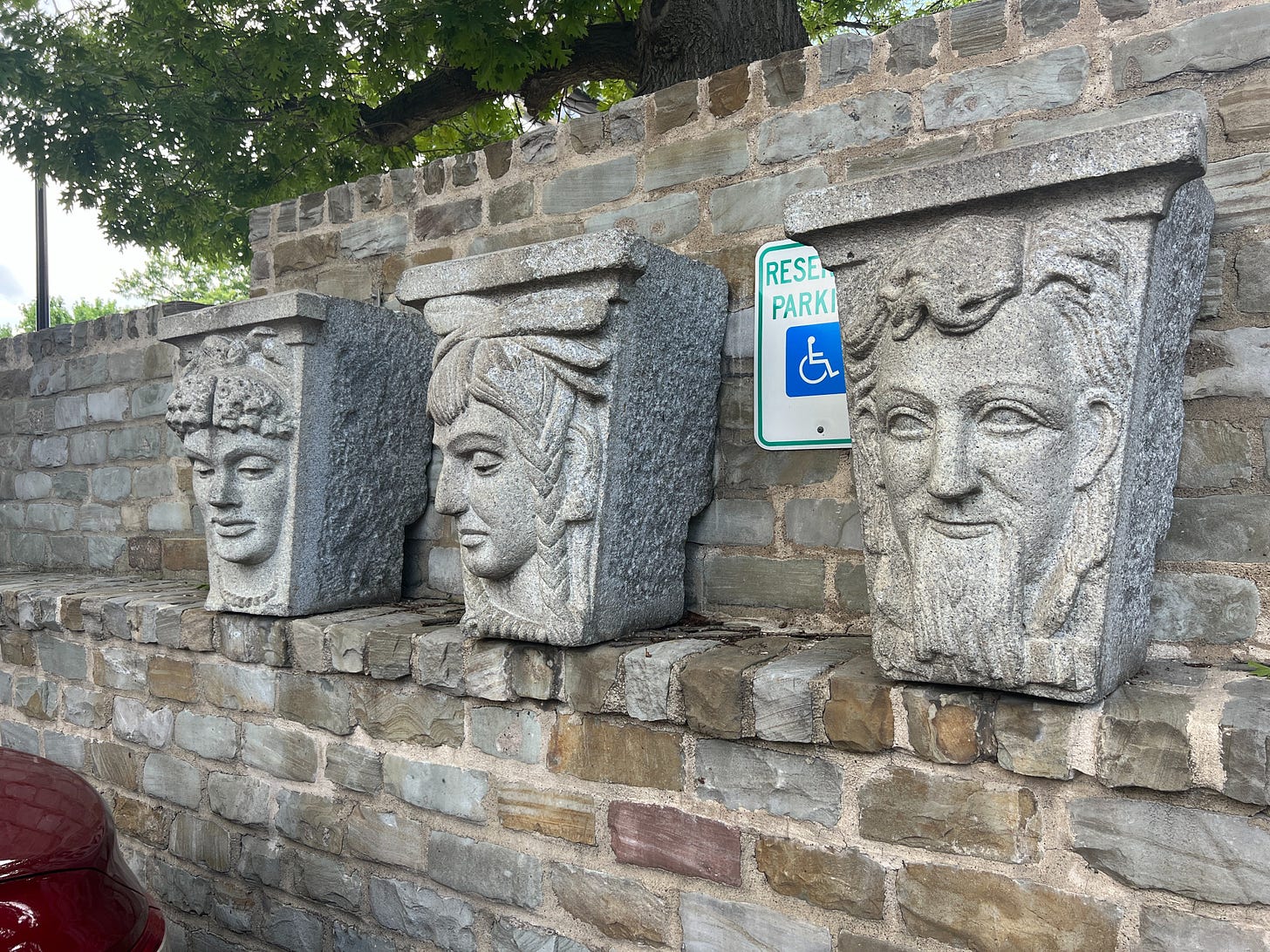 Three stone keystones with faces carved into them, resting on a brick shelf along an outdoor brick wall