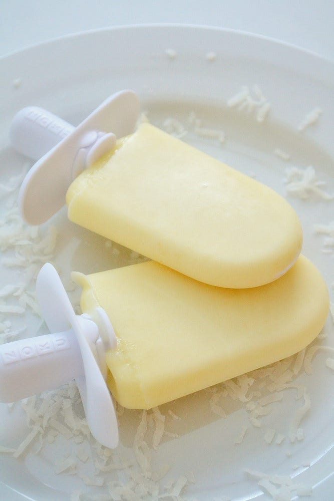 Two yellowish popsicls on a white plate sprinkled elegantly with coconut. "Pineapple Coconut Popsicles" by jamieanne is licensed under CC BY-ND 2.0. 