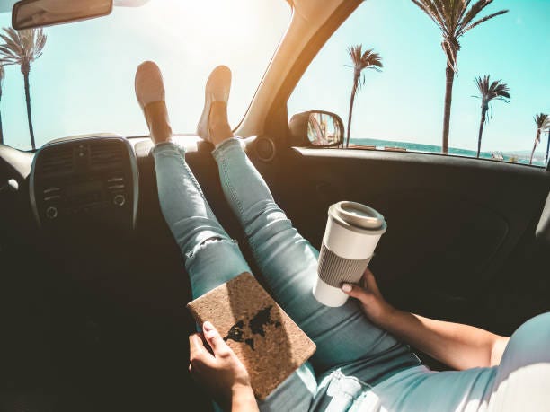 Woman drinking coffee paper cup inside car with feet on dashboard - Girl relaxing in auto trip reading travel book with ocean beach and palms in background - Traveler concept - Focus on hands Woman drinking coffee paper cup inside car with feet on dashboard - Girl relaxing in auto trip reading travel book with ocean beach and palms in background - Traveler concept - Focus on hands california road trip stock pictures, royalty-free photos & images