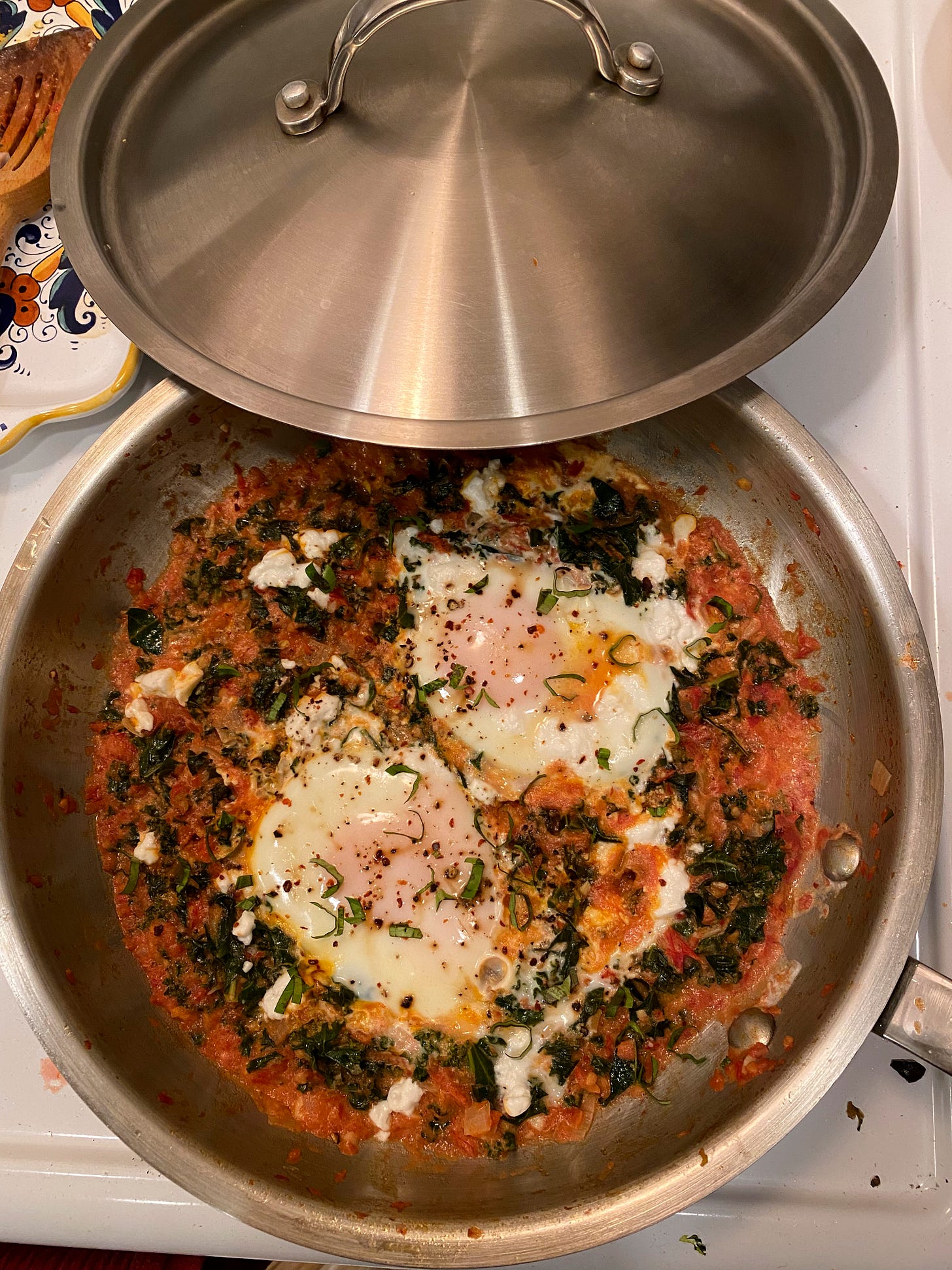 In a stainless steel pan on top of the stove, a creamy tomato sauce with pieces of kale. On top of it are two poached eggs and everything is sprinkled with chopped basil, pepper flakes, and crumbled feta. The lid to the pan is resting at the back edge.