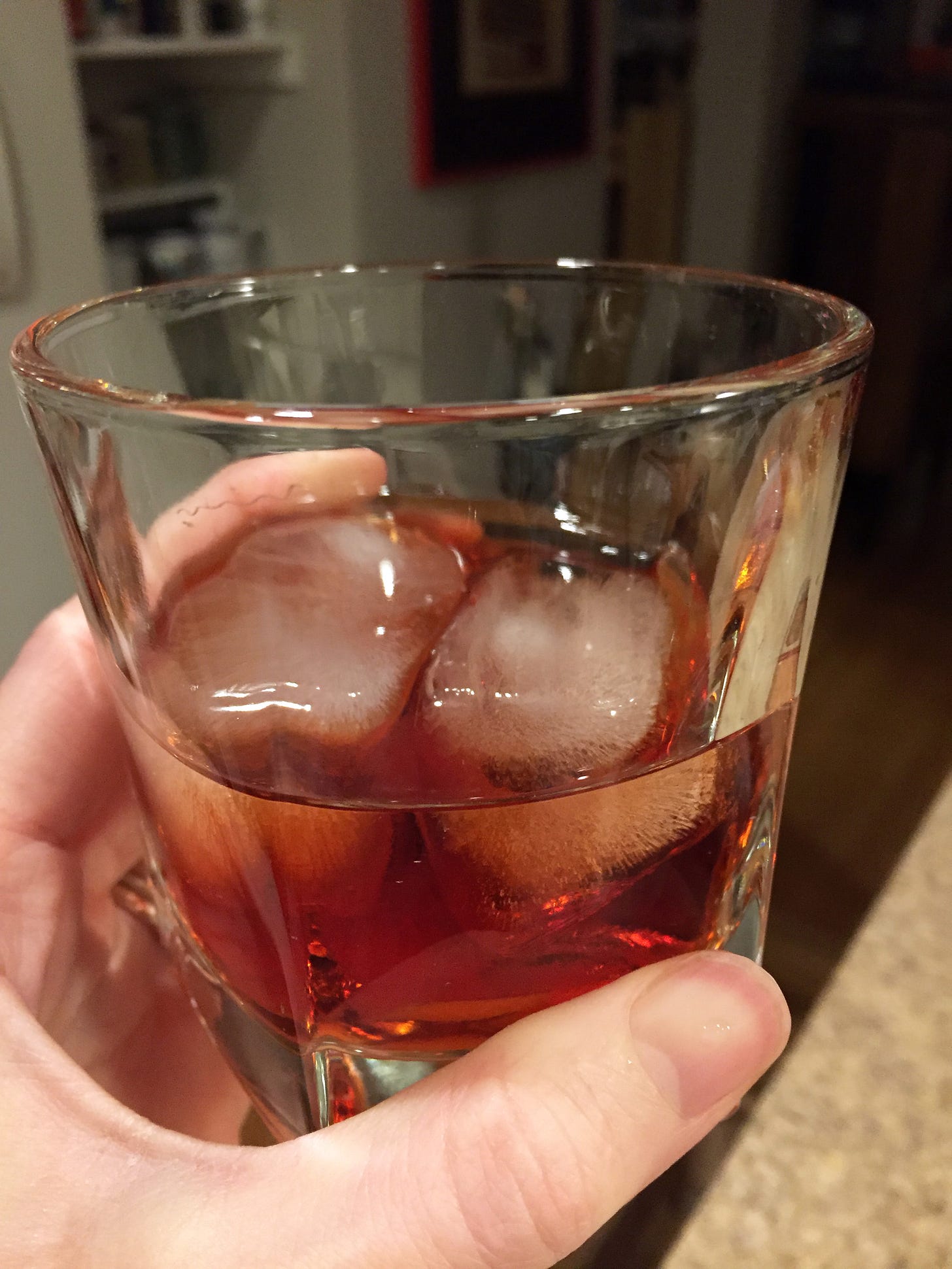 My hand holds a scotch glass about half-full with a red negroni with two ice cubes.