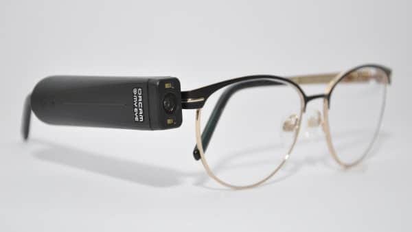 Foto do acessório para óculos MyEye Pro que transcreve em áudio o que está na sua frente. A armação é comum com os aros dourados e na lateral do lado direito possui uma caixinha preta cm uma câmera.