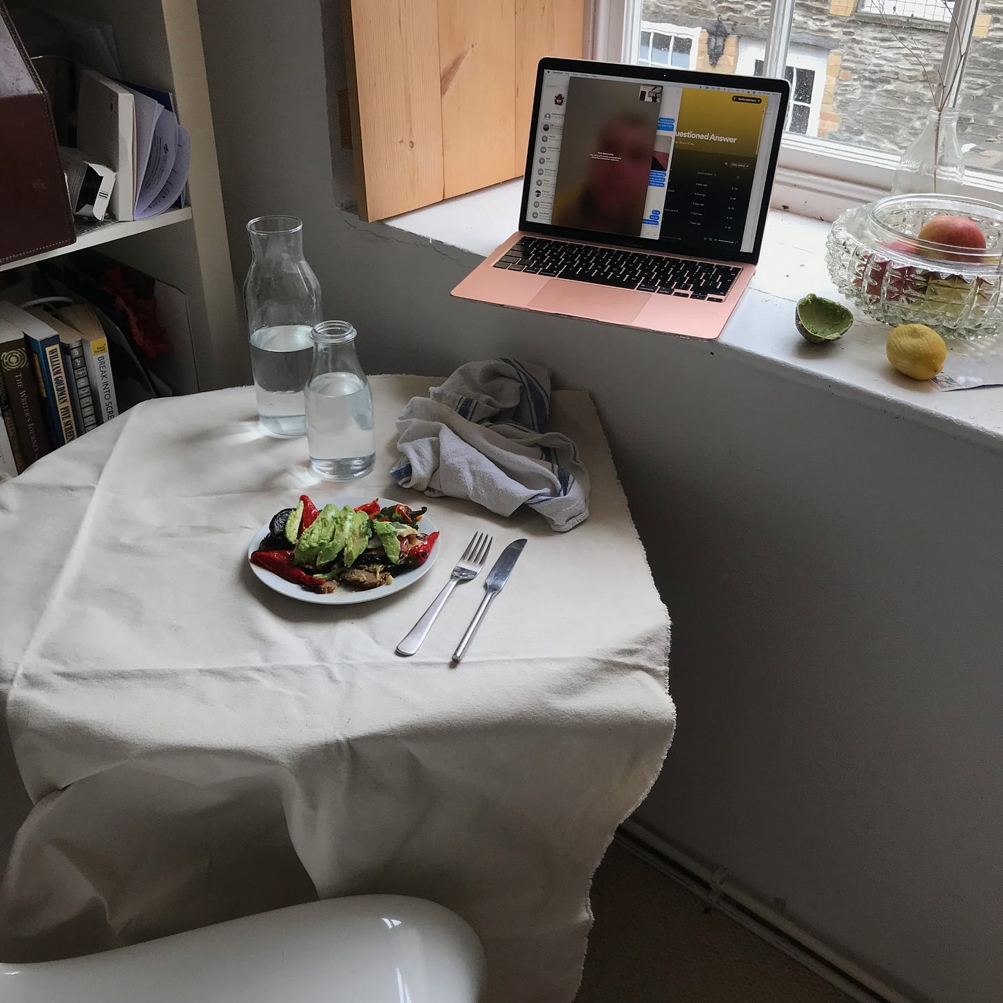 A shot of the same table, but from further back, later in the day. On the plate now are some ripe slices of avocado, more peppers, topping, jumbled over. In the background is a windowsill with a metallic pink laptop and half an avocado skin on it.  
