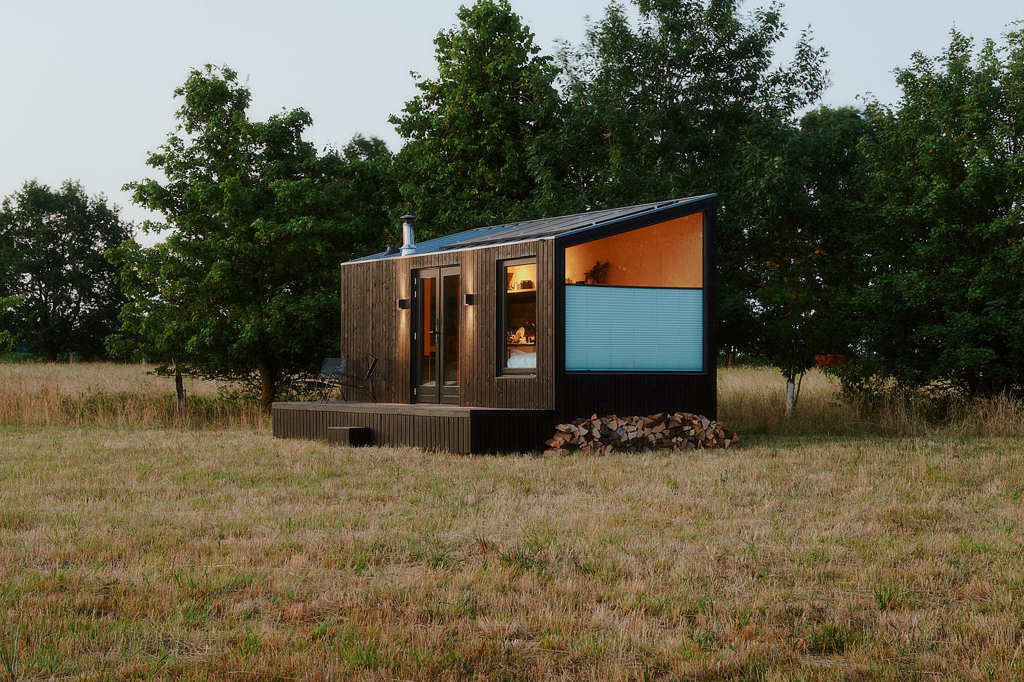 A modern black cabin with a sloped roof in a field, with trees behind.