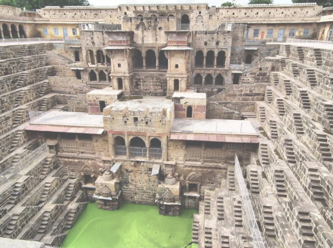 An outdoor picture of a large stepwell made from beige bricks designed with arches and carvings in geometric patterns that create many small descending steps all along the inner wall of the well that go all the way down to the water which is green colored.