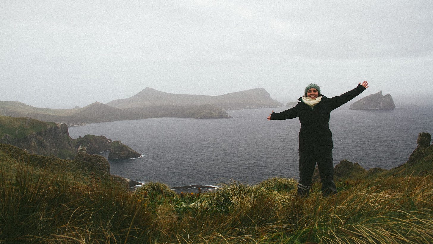 A photograph taken with a self-timer, with Louise standing with their arms out, looking at the camera - they are wearing a raincoat, long pants, a thick white scarf, and a green woollen headband. Behind them is a wide landscape looking out across two large bays and an island on the righthand side. More directly behind them is a harbour with cliffs going straight down to the ocean.