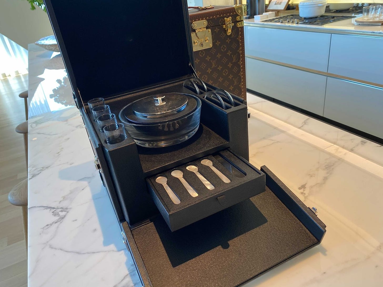 The caviar trunk is a matte black cube, top lid tilted up and front folded down to reveal a drawer of caviar spoons, and a bowl, presumably to hold the caviar. It sits on a shiny marble countertop and looks insanely wealthy.