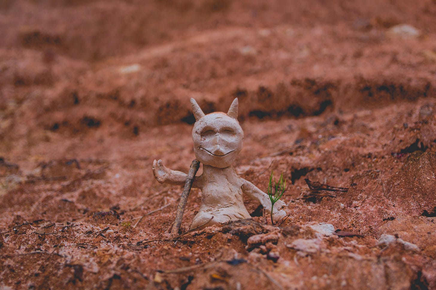 A small crude clay sculpture of a horned devil is posed on rocky red ground with a tiny twig leaning like a staff against its shoulder.