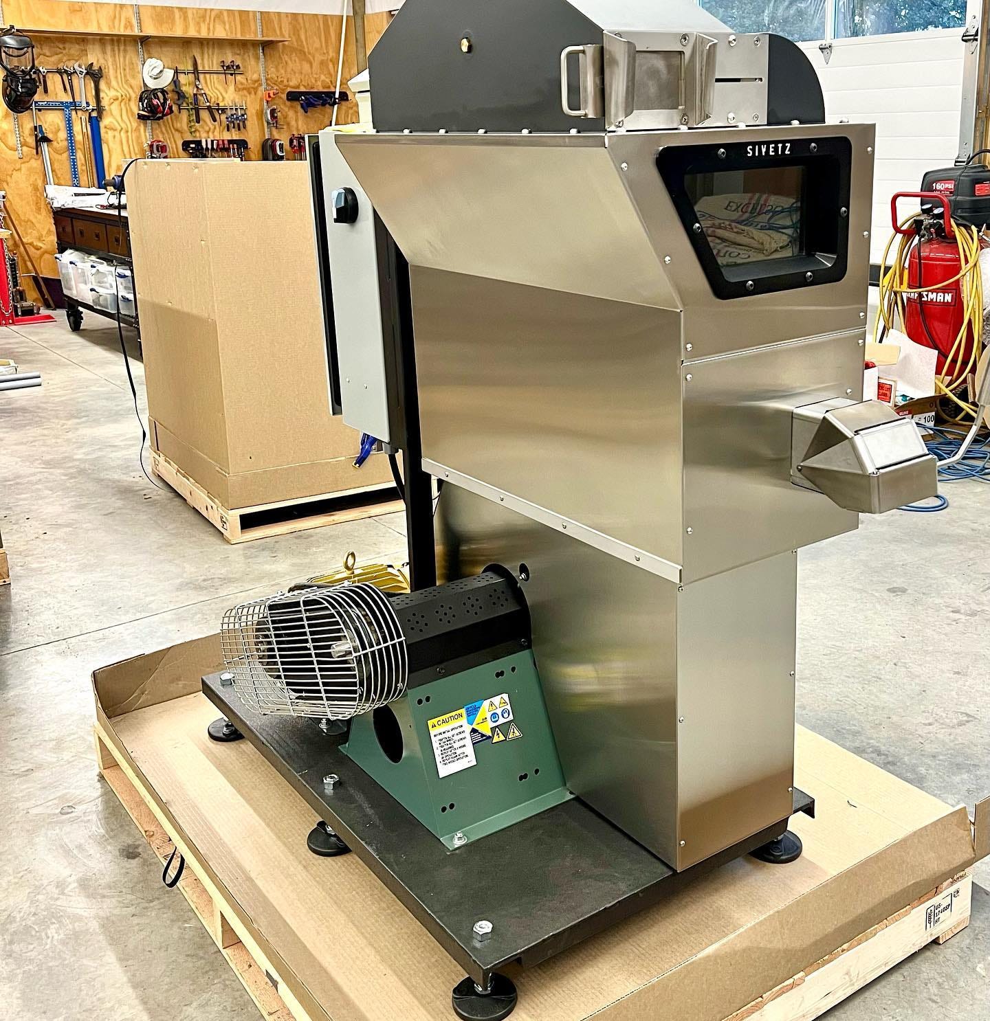 A stainless steel coffee roasting machine. Three rectangular steel boxes are stacked. The top one has an angled side and a window into the machine. A green