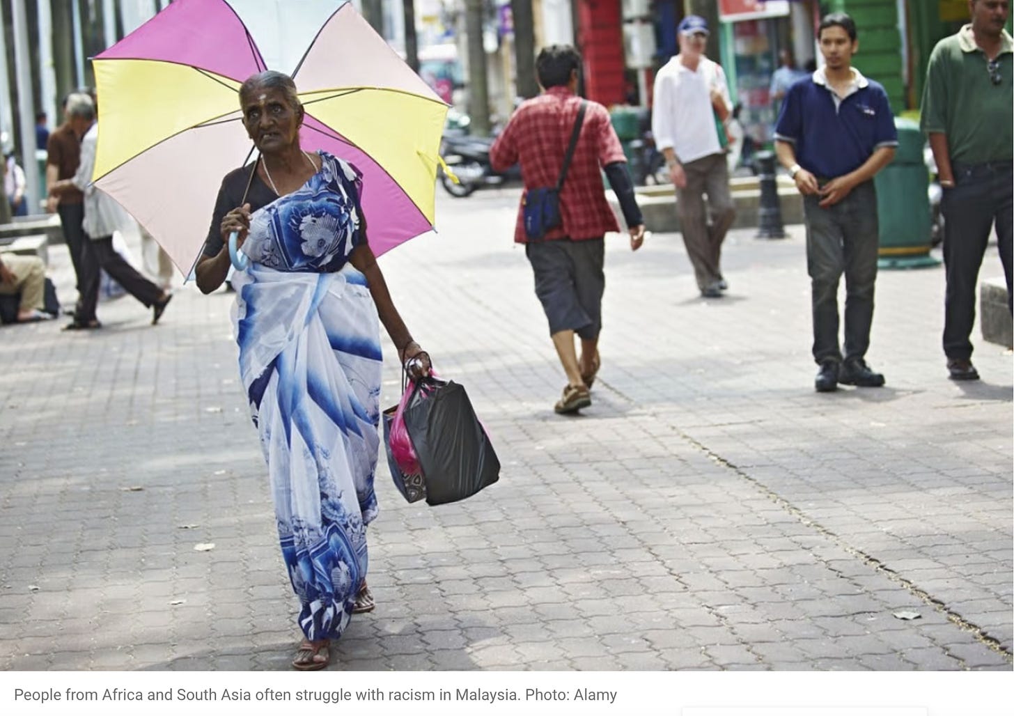 A picture of an older dark skinned woman wearing a white and blue sari and a bindi on her forehead. She is carrying an umbrella and some bags and walking alone down the street. Lighter skinned people walk around her or stand further away. Caption below picture reads: “People from Africa and South Asia often struggle with racism in Malaysia.”