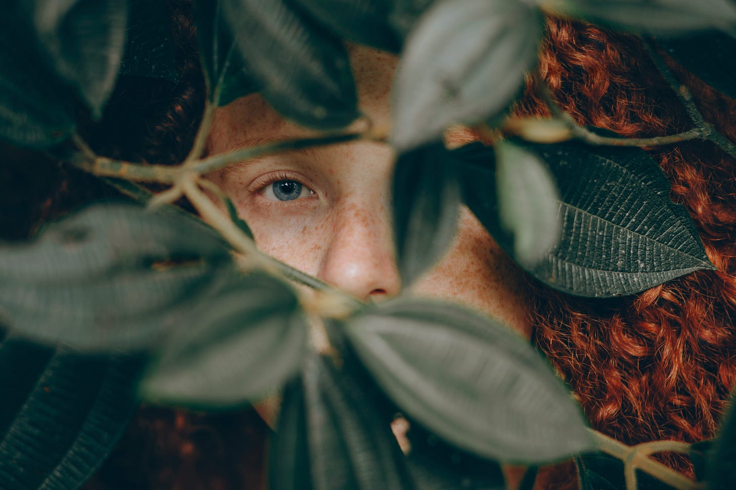 A red-haired person hiding behind green leaves.