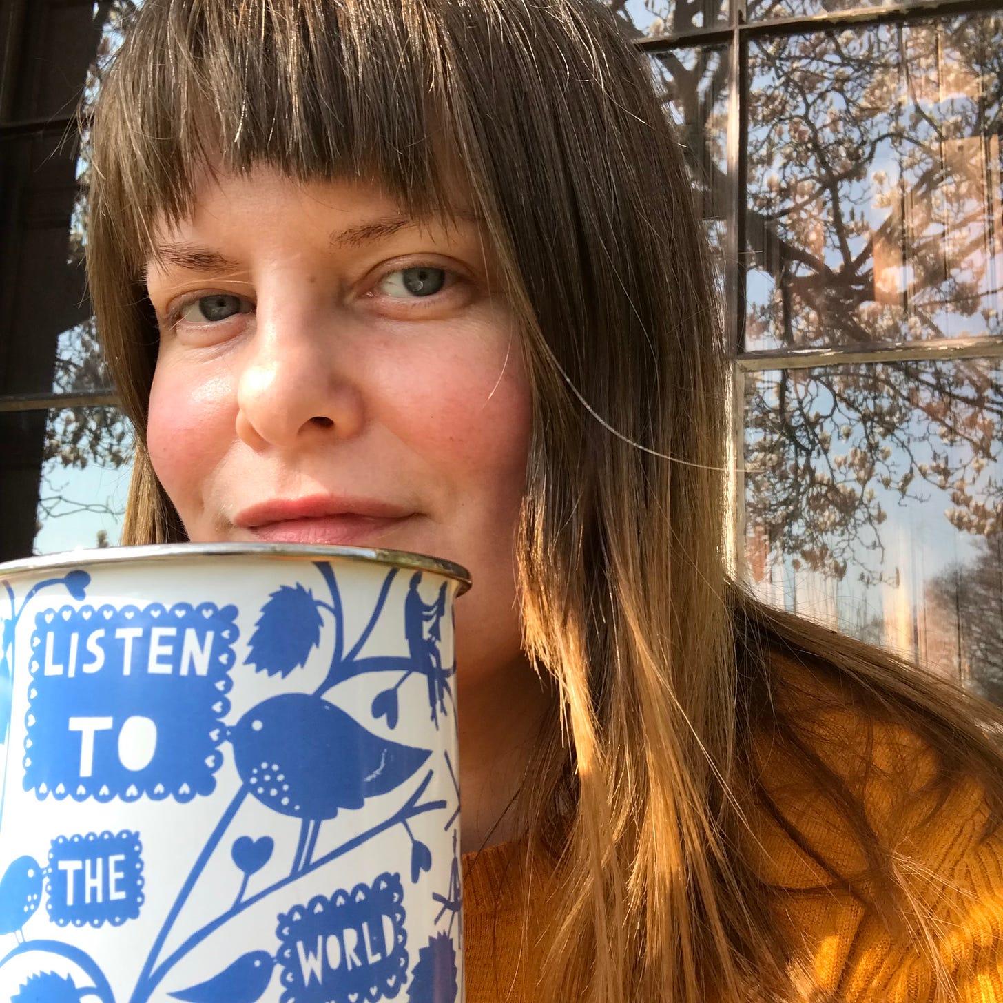Alice, a white woman with brown hair looks at the camera. She is holding an enamel mug with a blue pattern which says 'listen to the world'