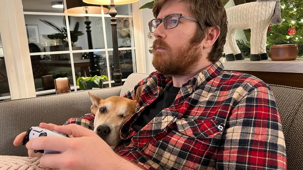 The author sits on the couch holding a PlayStation 5 controller with his right arm around his dog, who looks at the camera.
