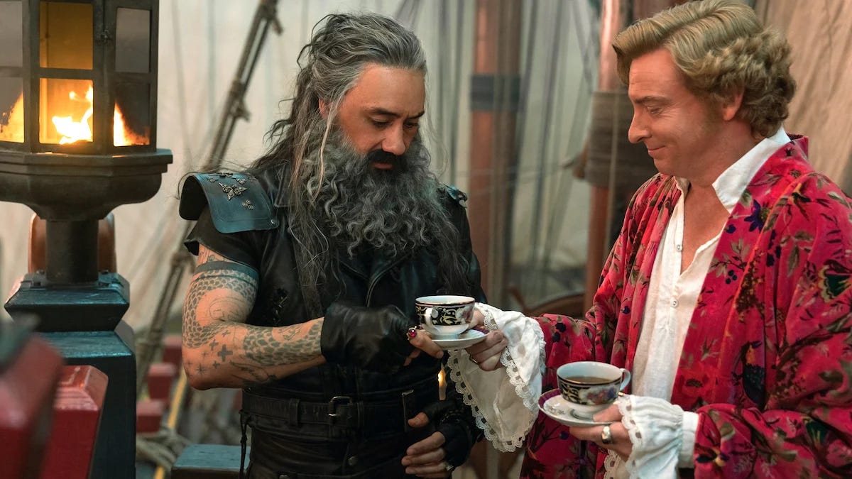 ed, a pirate wearing leather and sporting long messy hair and a beard is handed a cup of tea by stede, an elegant man with short curly blonde hair and wearing an elegantly decorated robe