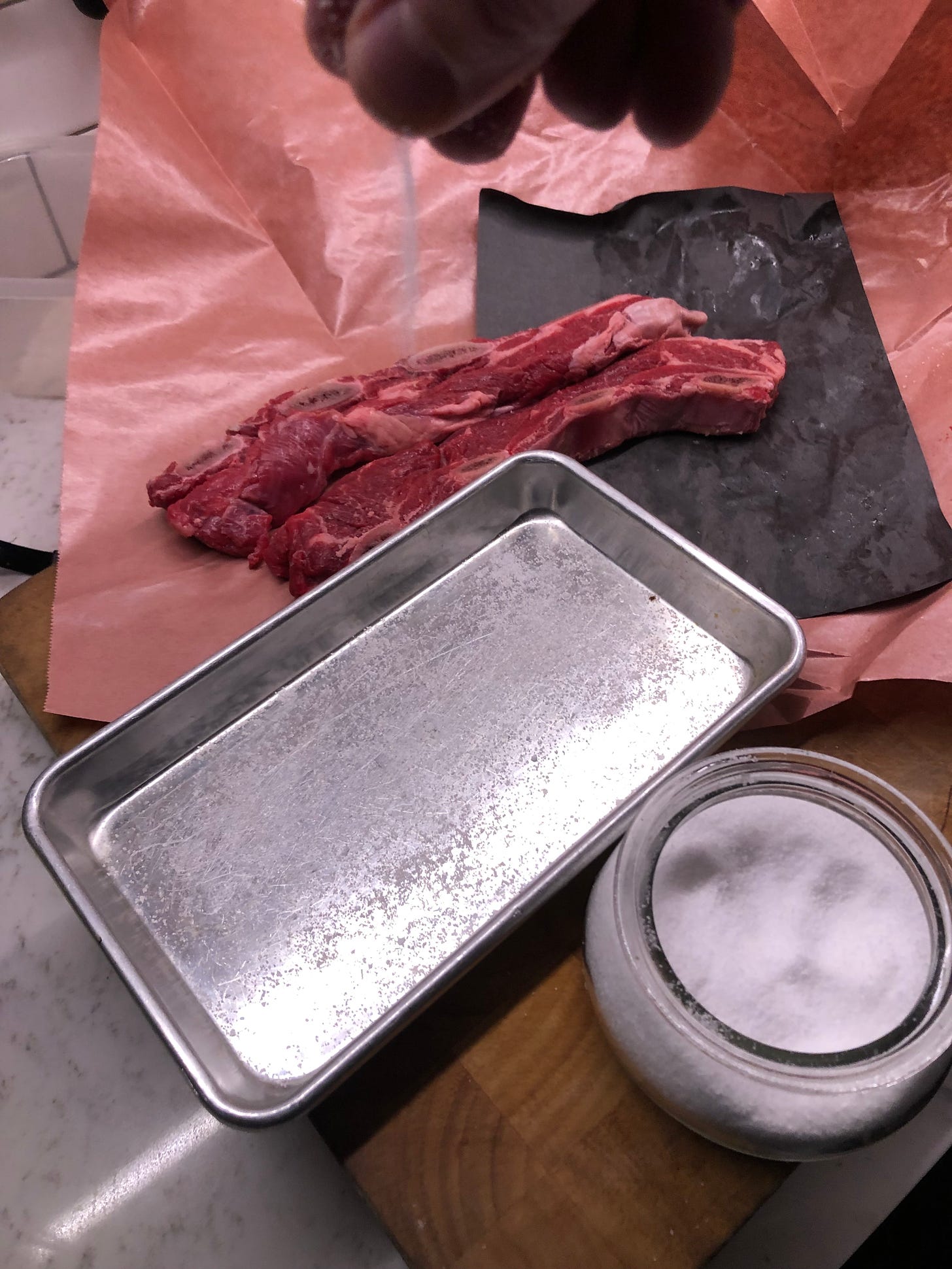 Two pieces of beef short rib are sitting on an opened package of butcher paper. A hand is sprinkling kosher salt onto a small sheet tray. 
