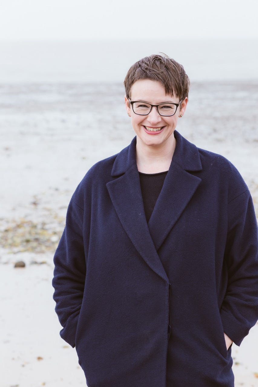 Katherine May standing in front of the sea and smiling at the camera