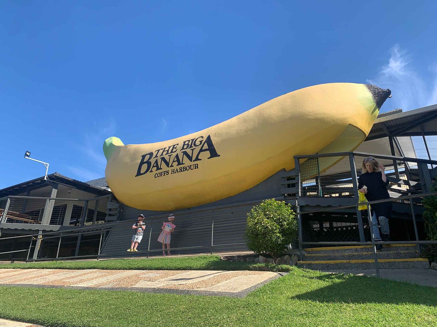 Two small children stand in front of The Big Banana at Coffs Harbour, under a bright blue sky.