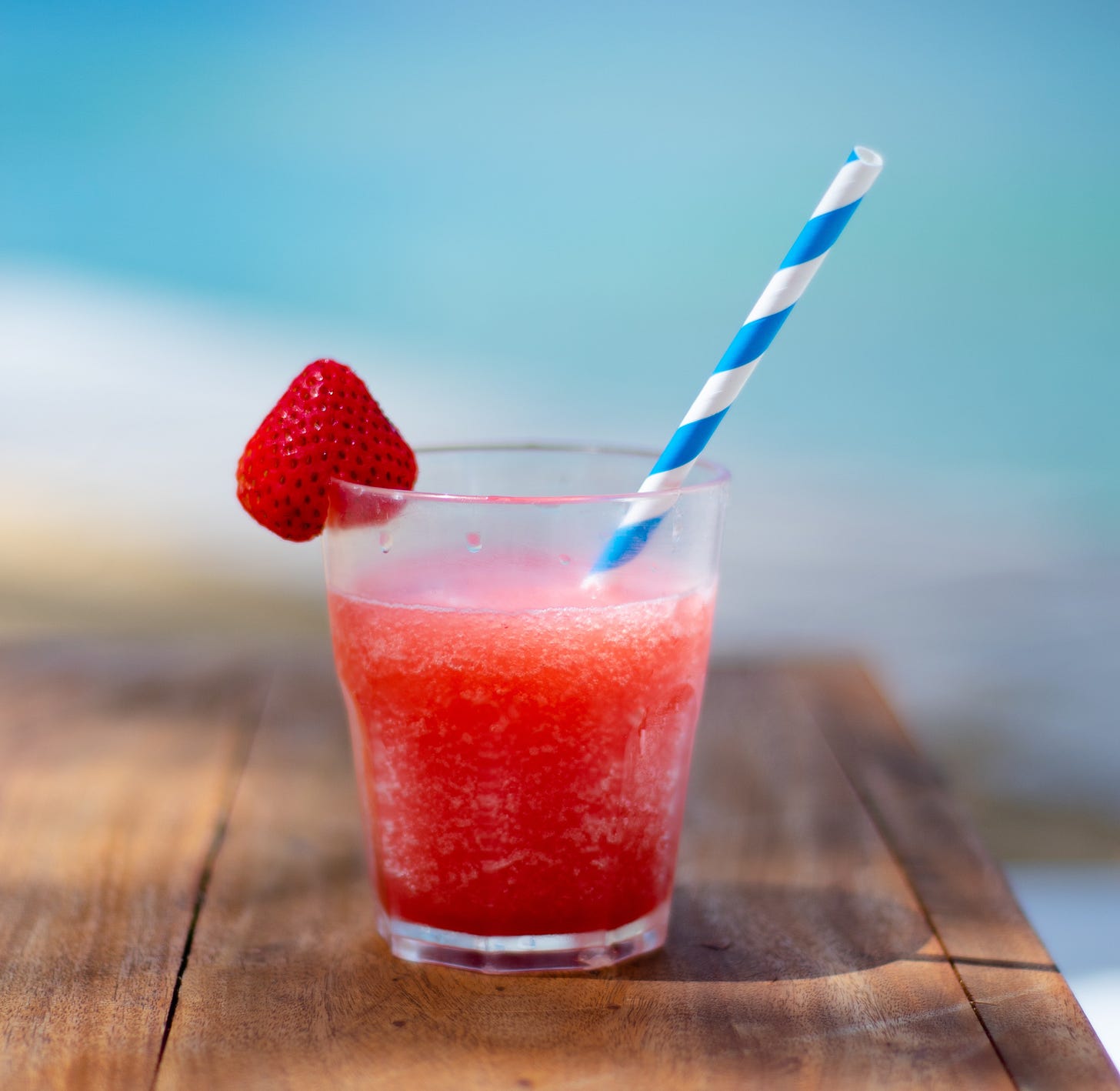 A pink drink with a strawberry wedged on the glass with a spiral white and blue straw.