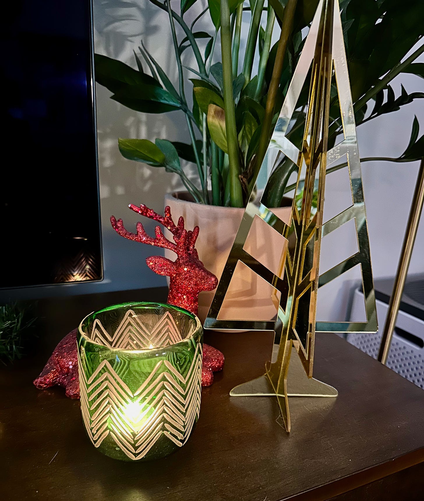 Green glass candle holder with tree pattern, gold lucite tree, and red glitter reindeer laying down with a ZZ plant in the background