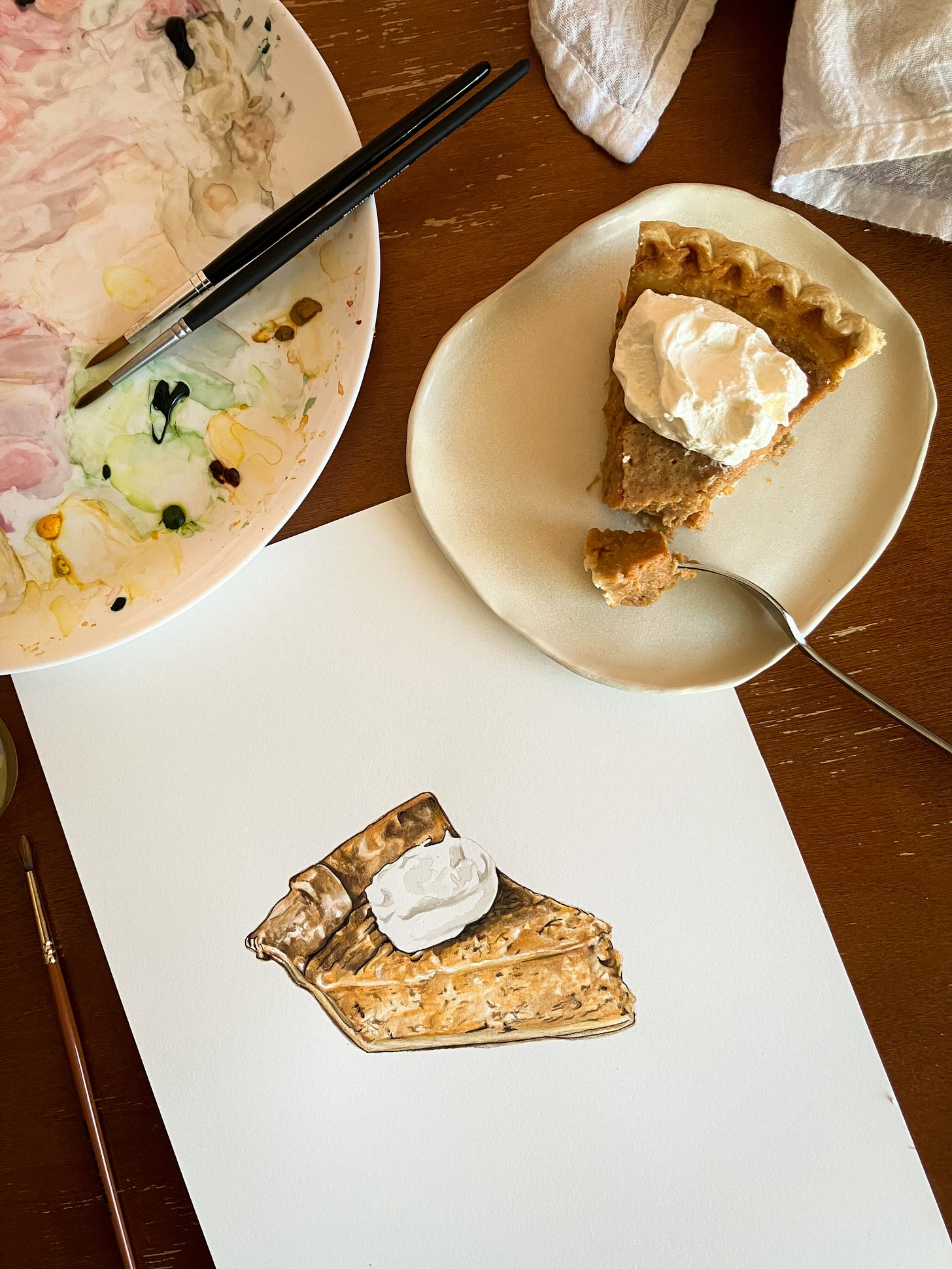 An overhead shot of a slice of pie next to the painting of the slice of pie with the watercolor palette close by 