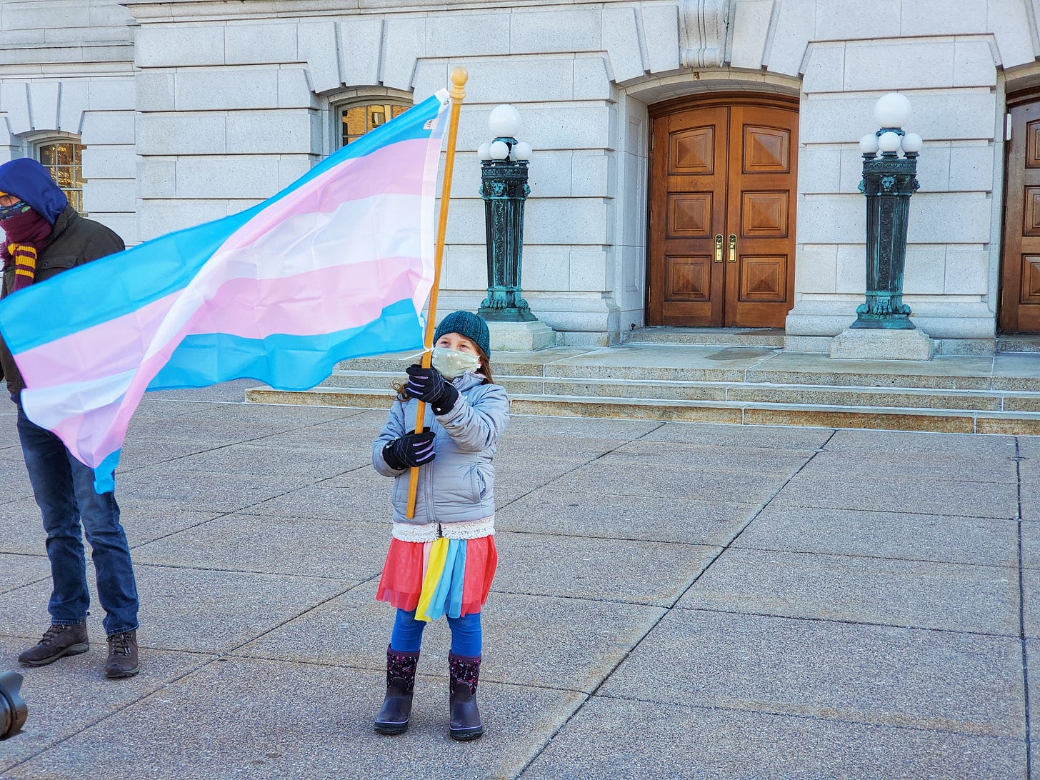 A young child waves a large transgender pride flag. She's wearing a knit cap, face mask, winter coat, and bright rainbow skirt over leggings.