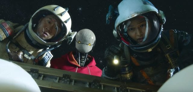 Screencap of a scene from Space Sweepers, on Netflix. Three faces -- two human, one robot -- peer into an outerspace storage container they've just opened. There's a surprise inside!