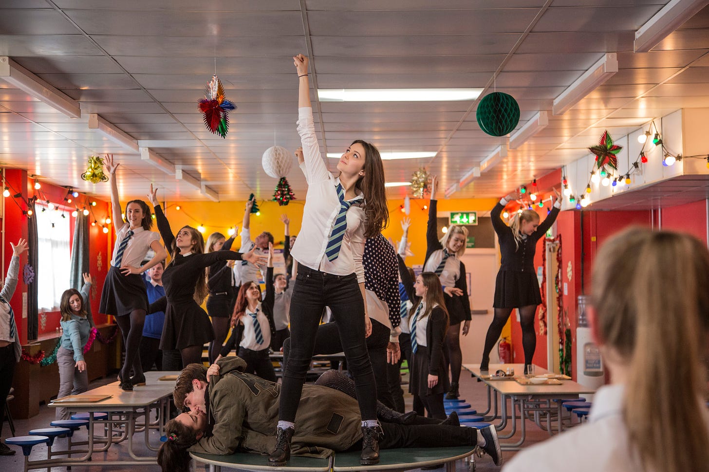 Still from Anna and the Apocalypse. A white girl with long brown hair and wearing school uniform stands on a table, arm raised. Her classmates are behind her, also with arms raised, mid dance routine.