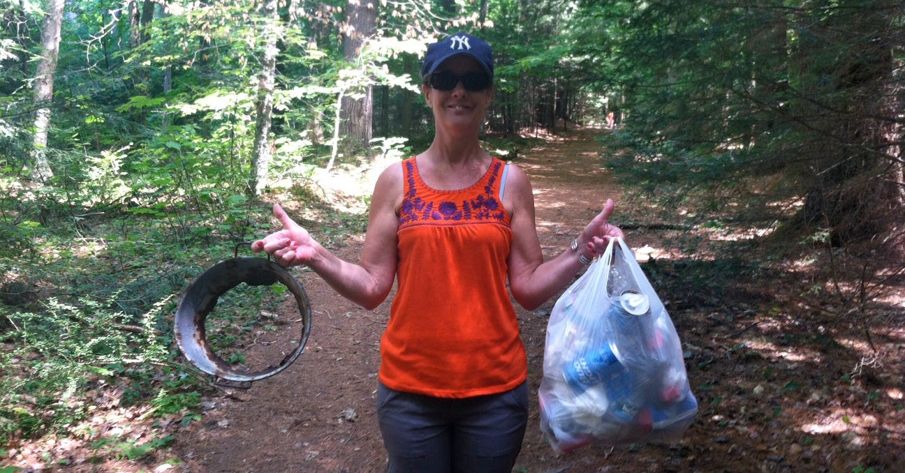 My friend Mary holding a rusty piece of metal and a bag of trash on a ndddle strewn Adirondack path.