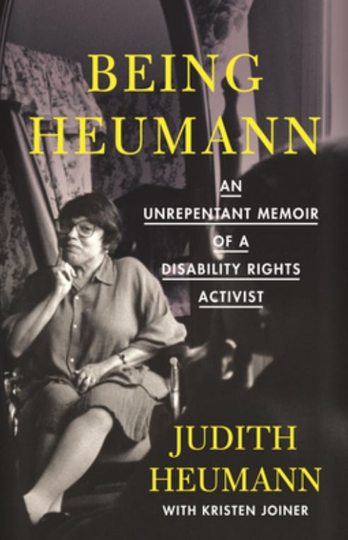 Book cover of Being Heumann: An Unrepentant Memoir of a Disability Rights Activist, by Judith Heumann with Kristen Joiner. Cover photo shows a smartly dressed middle-aged woman with brown hair and glasses wearing a skirt and collared shirt sitting in a wheelchair with crossed legs. She has a knowing smile on her face and her one hand is resting below her chin.
