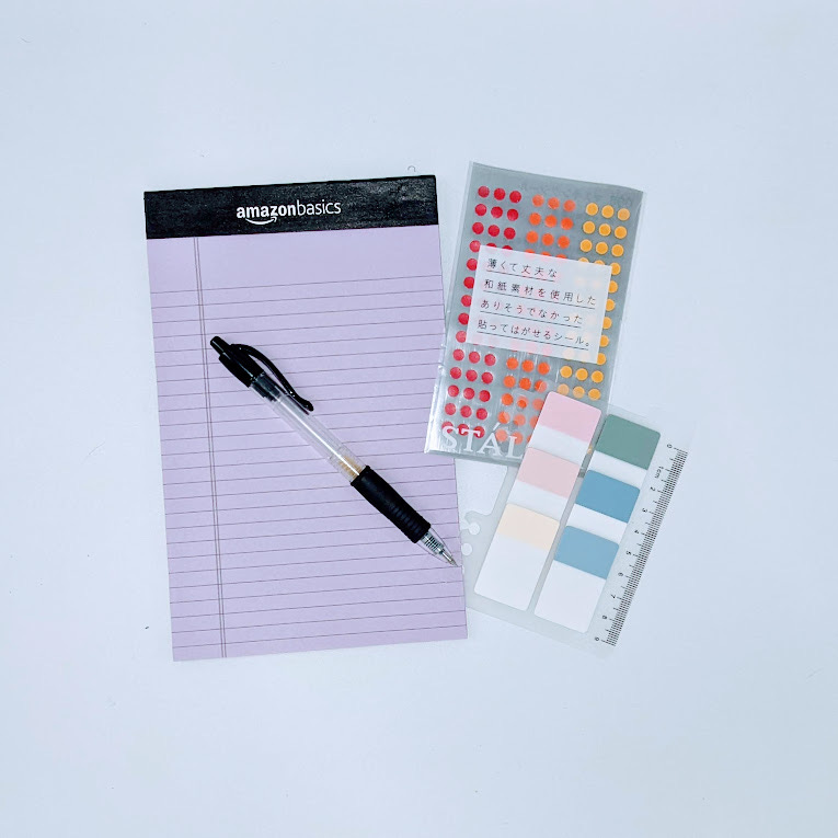 A black pen sitting atop an AmazonBasics notepad with lined, purple paper; a pack of small dot stickers in red, orange, and yellow with Japanese writing on the package; and a pack of sticky tabs in shades of pink, yellow, and blue.