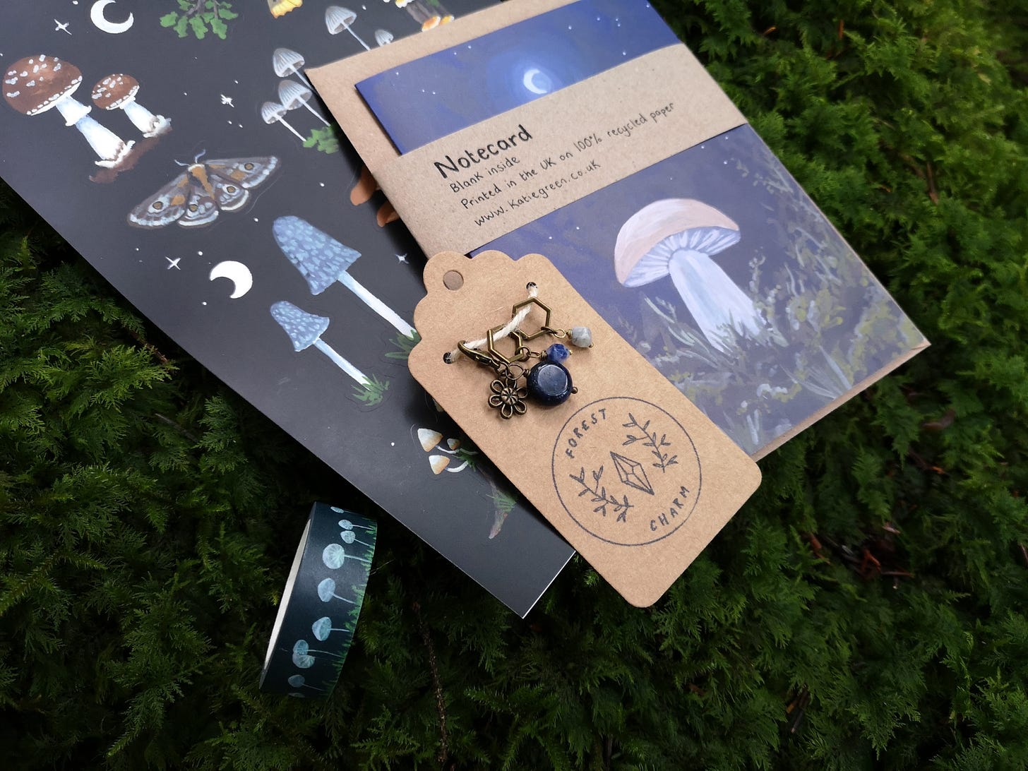Image description: A bundle of goodies lying on some squishy green moss: a set of stitch markers and progress keepers featuring sodalite beads and a flower charm, lying on top of a notecard with a mushroom and moon, some stickers of mushrooms moths and moons and a roll of dark blue washi tape with white mushrooms.