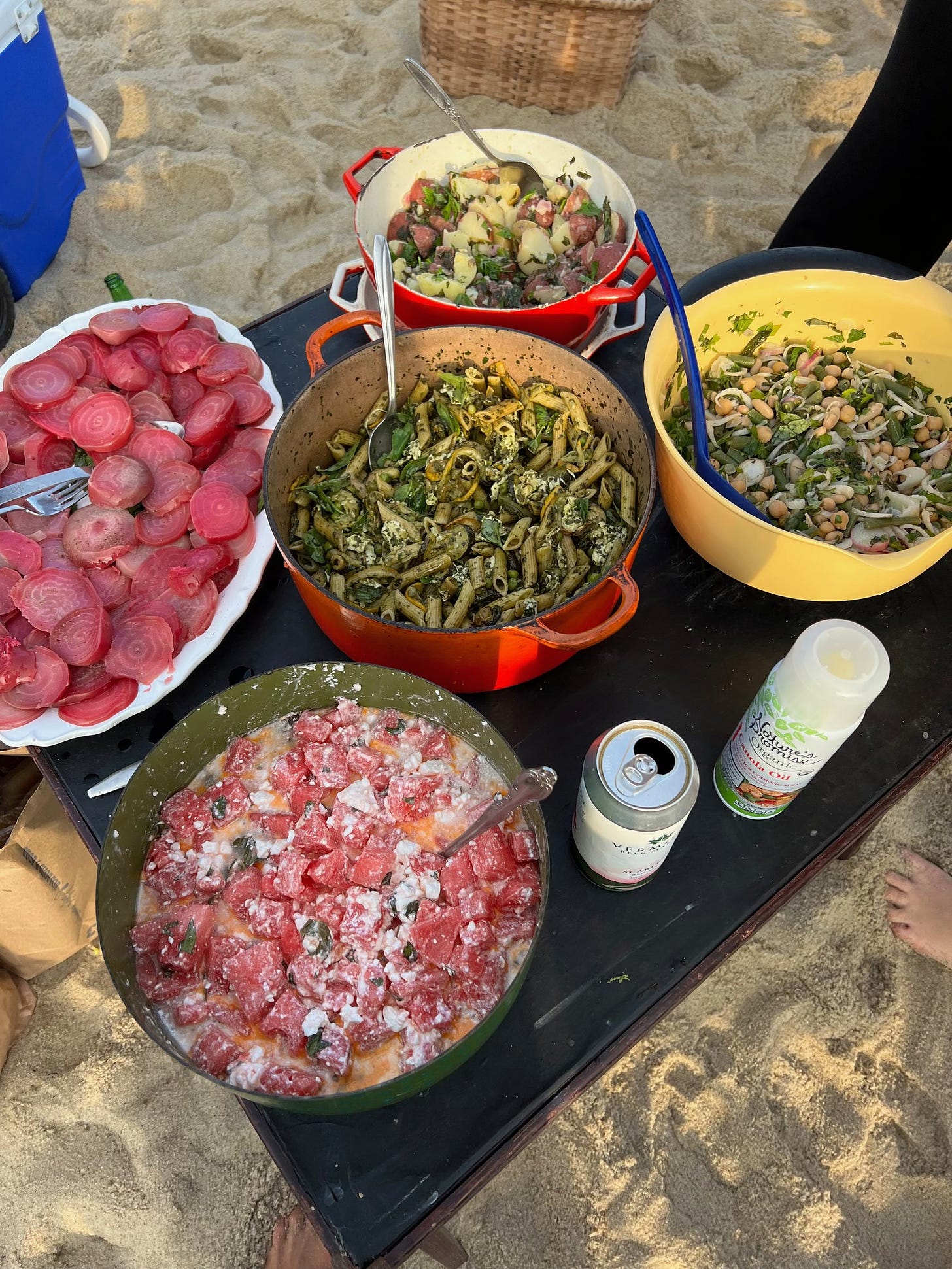 A card table on the beach holding five large bowls of salad: a platter of sliced beets, a pasta salad, a potato salad, a three bean salad, and a watermelon salad.