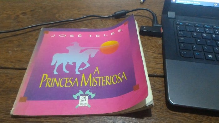 Livro “A Princesa Misteriosa”, de José Teles, com capa de fundo rosa e sombra de caveleiro montado em um cavalo com armadura e justa. O livro está em cima de uma mesa de madeira com notebook ao lado.