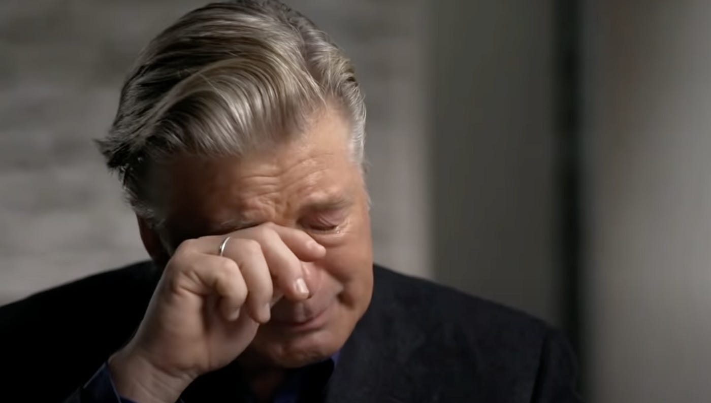 Image description: Alec Baldwin is wearing a black suit with a dark blue shirt, his grey hair combed back and his right hand raised to wipe a tear from his eye as he sobs. Behind him is a blurred grey background.