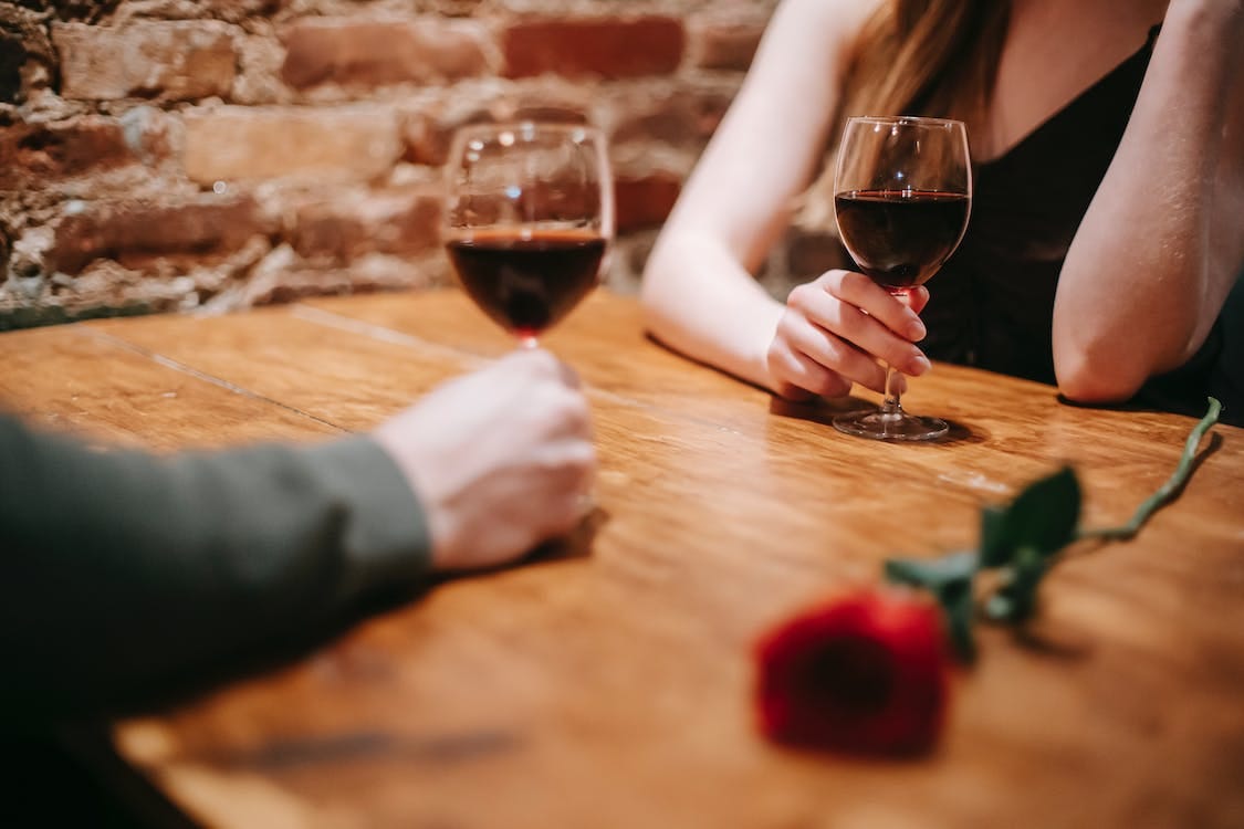 Free Crop anonymous couple in elegant outfits enjoying romantic date in bar at table with red rose and glasses with wine near brick wall Stock Photo