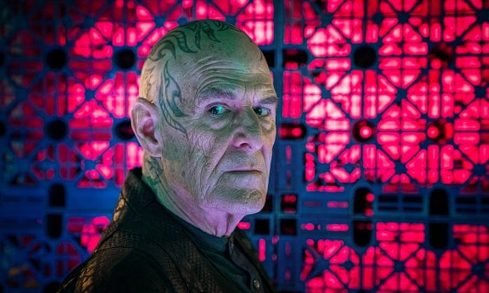 This image shows the character of Zellin (played by Ian Gelder) looking at camera with menace in his eyes. Tattoos cover Zellin's bald head. He stands in front of a red-and-purple wall covered with latches and crossbeams. This image shows the character of Zellin (played by Ian Gelder) looking at camera with menace in his eyes. Tattoos cover Zellin's bald head. He stands in front of a red-and-purple wall covered with latches and crossbeams.