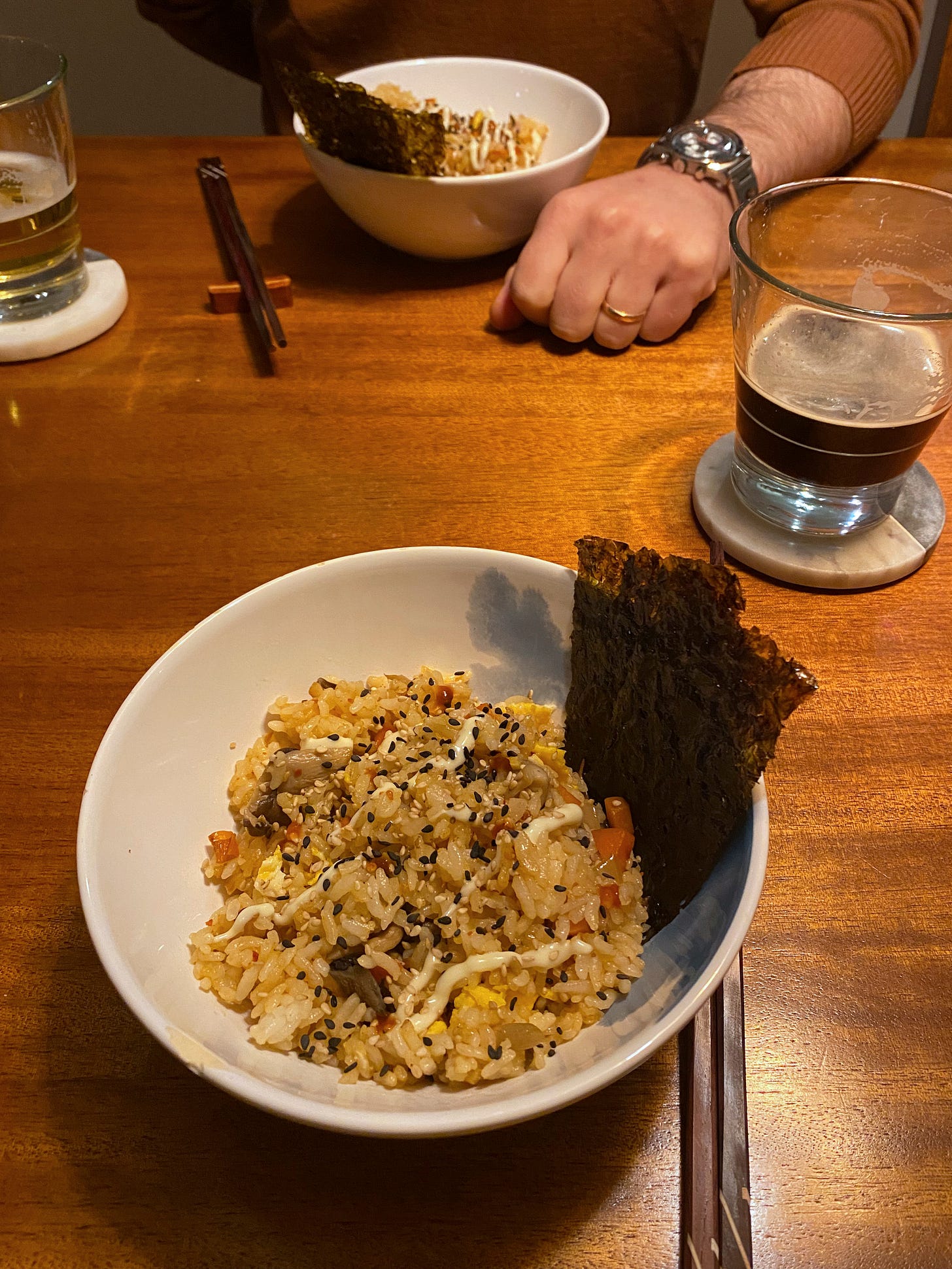 Across from each other on the table are two bowls of fried rice, each with pieces of gim at the side and a light drizzle of kewpie mayo and sriracha over top. Next to each plate is a set of chopsticks, and a half-full glass of beer on a stone coaster.