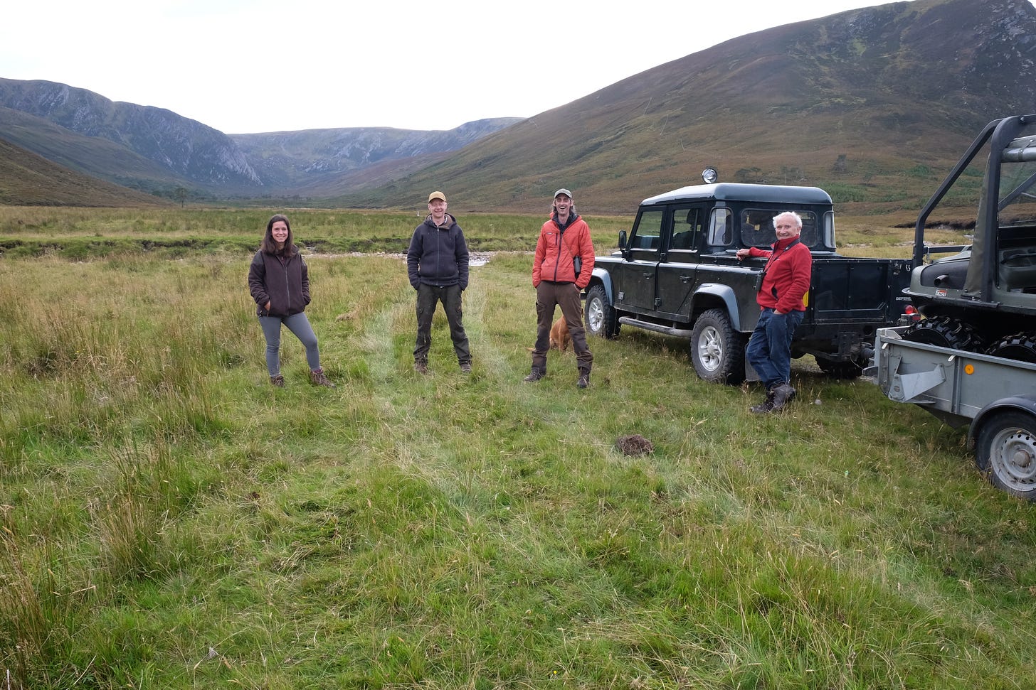 Three men and one woman stand in an open green space with Scottish vales in the background. The man on the far right is leaning against a land rover,