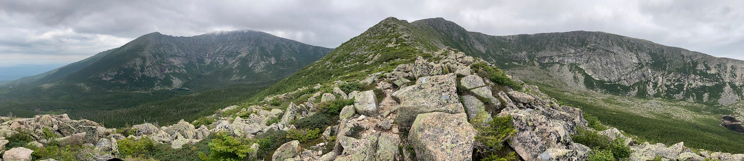 A panorama of Katahdin and Hamlin peak taken from midway up Hamlin ridge, about 3640 ft elevation, facing west. On the left are Pamola peak and Baxter peak above the South Basin, with the knife edge connecting them. In the middle is the spine of Hamlin ridge leading to Hamlin peak at center, and to the right is the North Basin surrounded by its wall of cliffs. The clouds are low and heavy, and the air is hazy from West Coast wildfire smoke. 