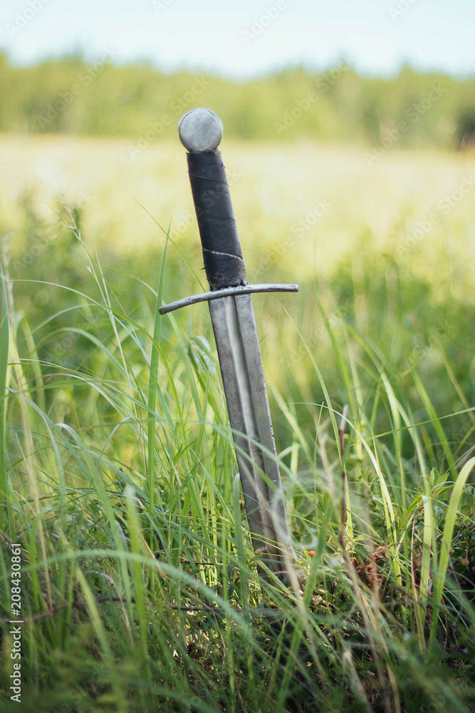 Knight's sword thrust into the ground. In the field against the background  of the forest and the blue sky. A sunny day of yellow-green steppe.  Battlefield. After battle. Crusade, medieval weapons. Stock