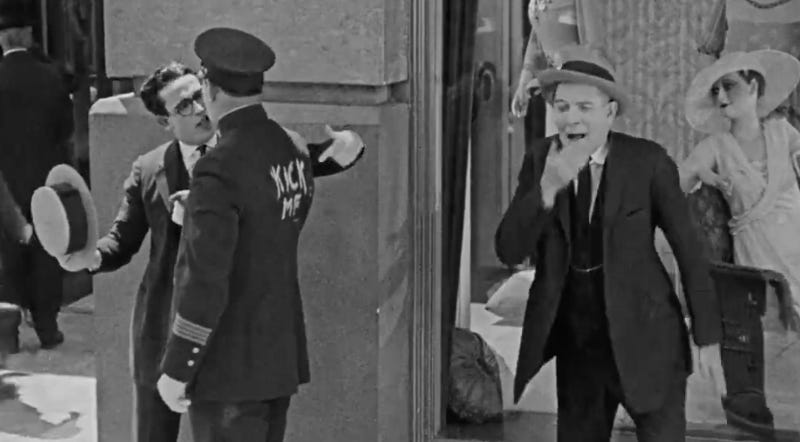 A man talking to a police officer, pointing at the words “kick me” written on his back while another man observes and laughs