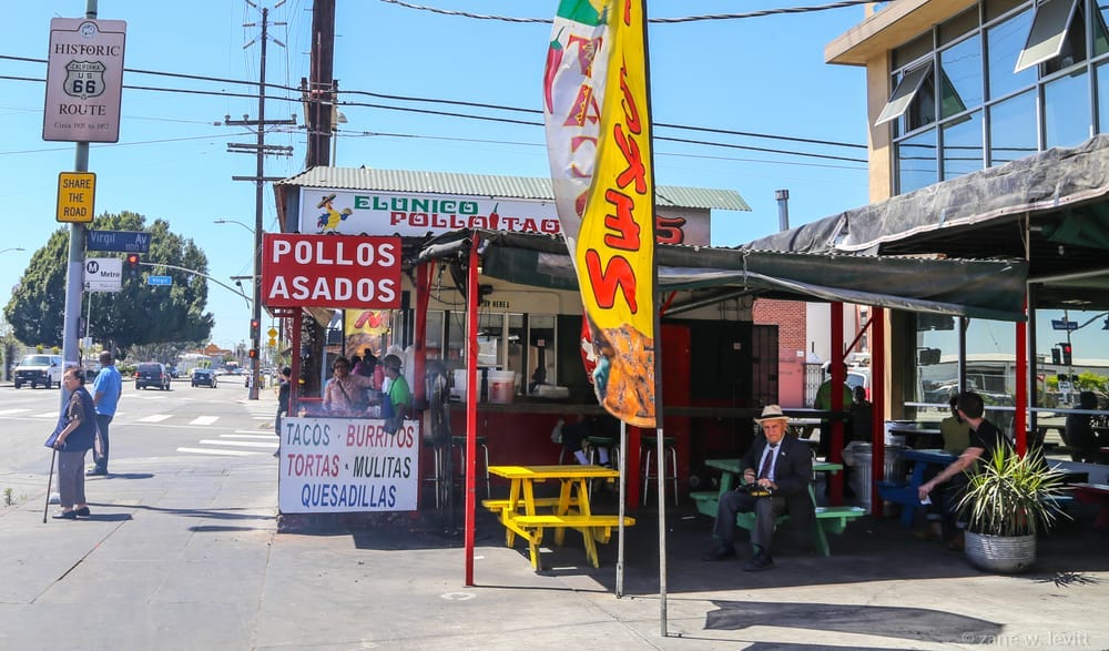 Shack on a corner with a red sign that says Pollos Asados in white and other signage indicating the sale of tacos, meats, etc. Yellow, green, and blue picnic tables sit under a metal awning. An older man sits at one of the tables, and a younger man sits at a different table. There are several other people around the premises and smoke is gathering near the grilled chickens. 