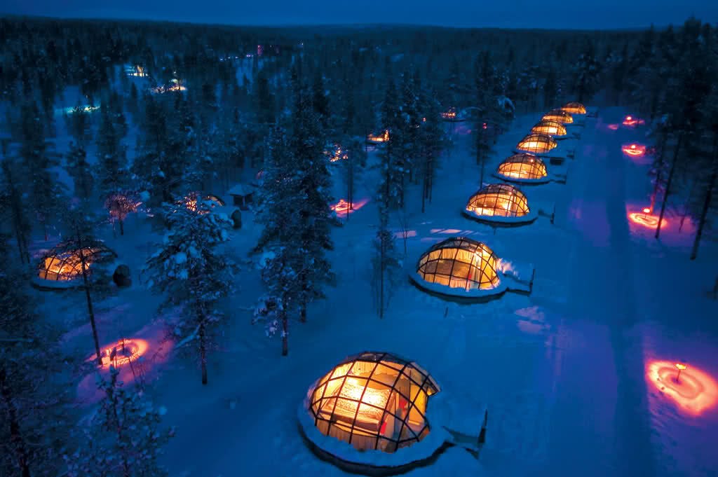 Two long rows of reinforced glass igloos, half buried in snow and illuminated orange from with, glowing in the purple dusk of a Nordic forest