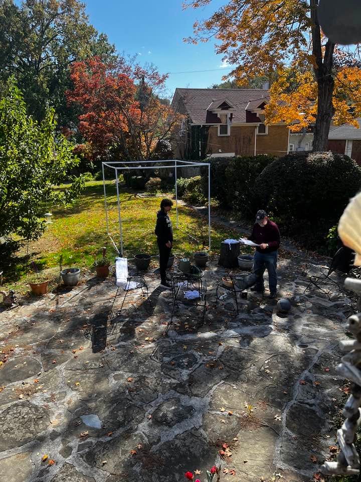 A white teenage boy and a middle aged white man construct the skeleton of a sukkah in a yard with lots of fall foliage.