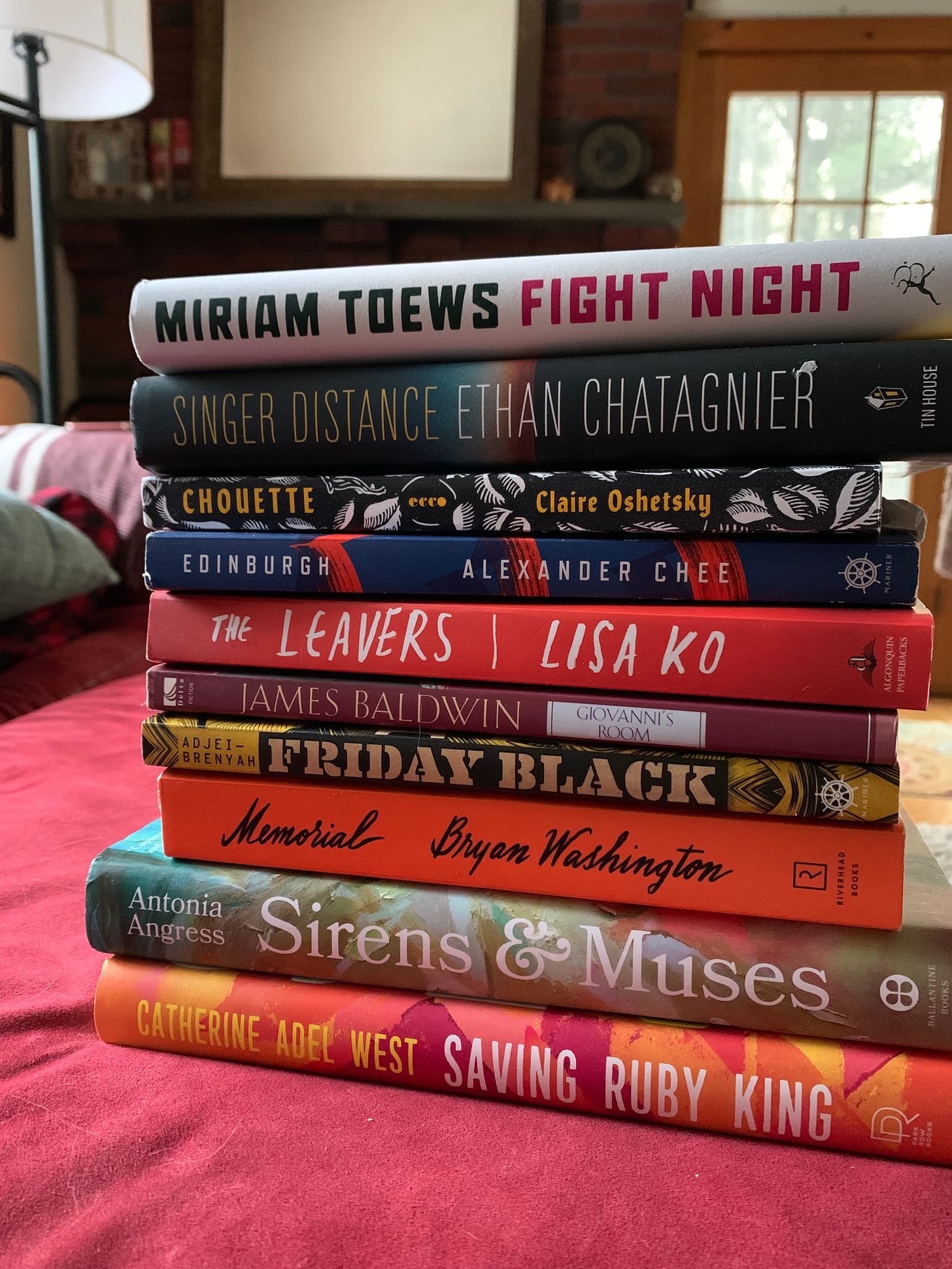 A stack of books on a red ottoman. Books from top to bottom include FIGHT NIGHT by Miriam Toews, SINGER DISTANCE by Ethan Chatagnier, CHOUETTE by Claire Oshetsky, EDINBURGH by Alexander Chee, THE LEAVERS by Lisa Ko, GIOVANNI'S ROOM by James Baldwin, FRIDAY BLACK by Nan Kwame Adjei-Brenyah, MEMORIAL by Bryan Washington, SIRENS & MUSES by Antonia Angress, and SAVING RUBY KING by Catherine Adel West