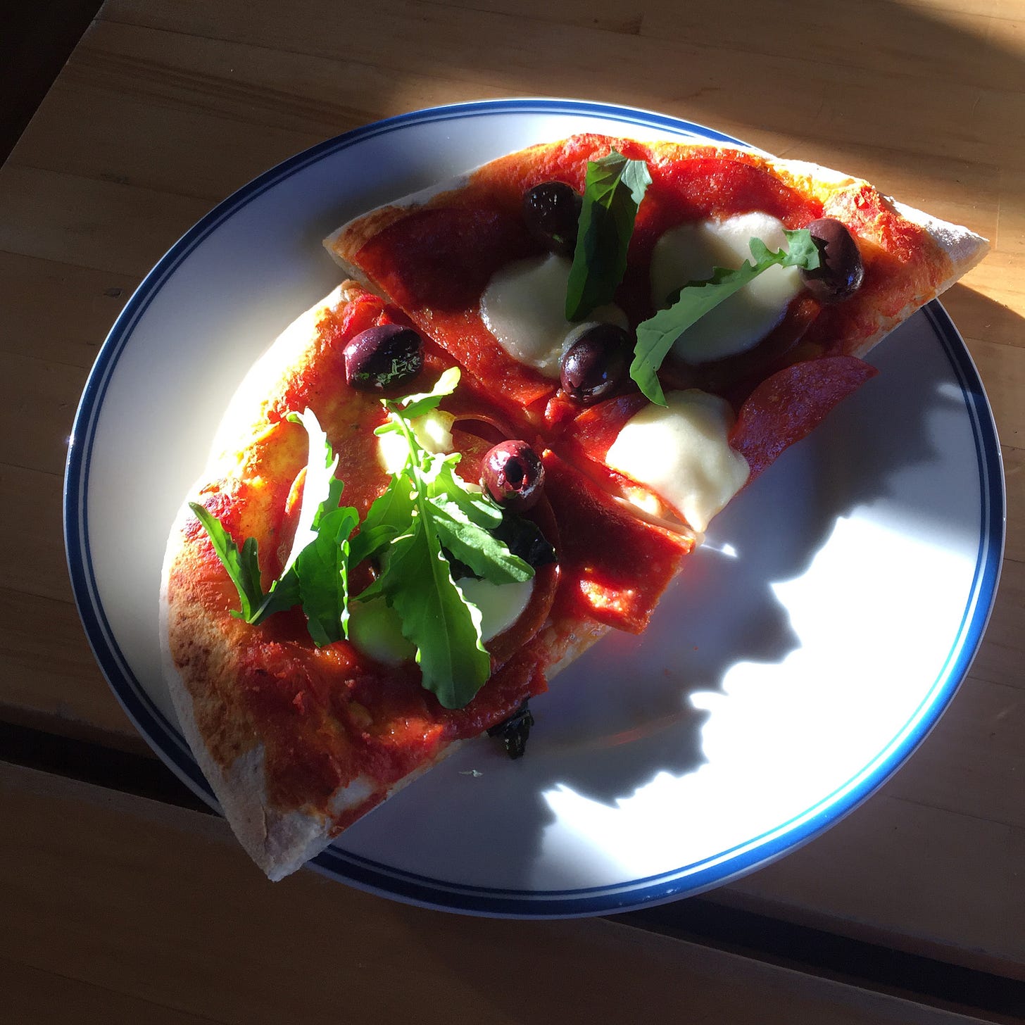 two slices of pizza with salami, kalamata olives, and slices of bocconcini with fresh arugula on top. They sit on a white plate, and dappled light from a window creates shadows across the plate and table.