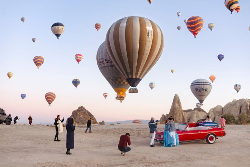 Il cielo della Cappadocia è pieno di mongolfiere, mentre ai piedi ci sono diversi turisti intenti a fotografare la scena.