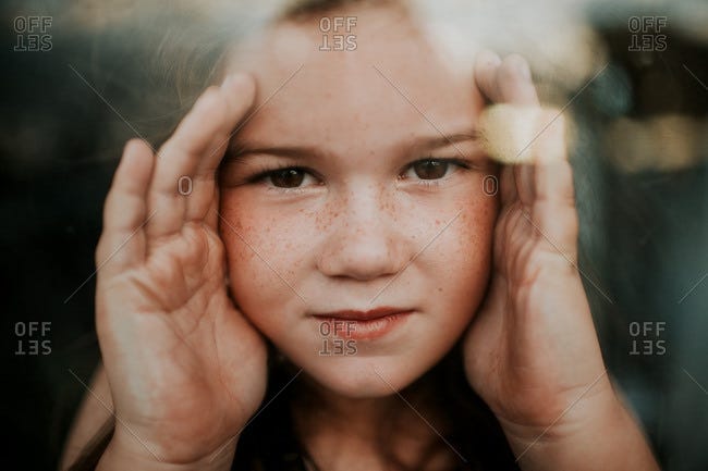 Girl with brown eyes and freckles with her hands up against glass window  stock photo - OFFSET