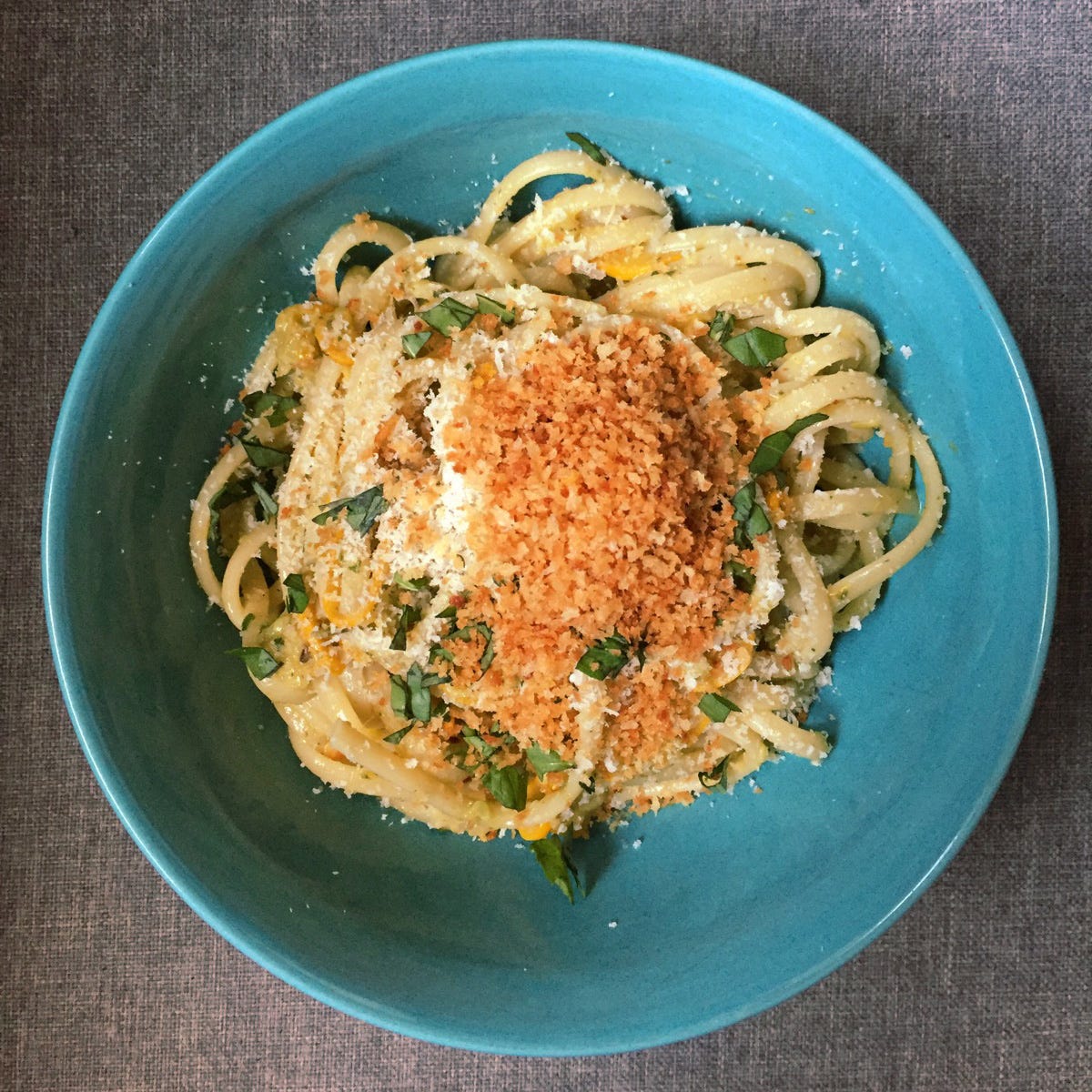 a blue bowl of linguine with slices of yellow summer squash, shreds of basil, parmesan, and toasted panko crumbs on top.