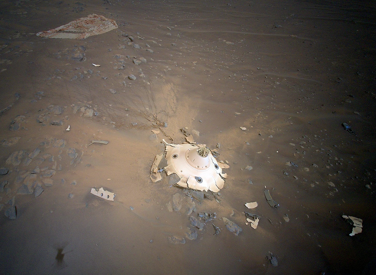 The pale, smashed backshell of the Persverance rover lies on the red-brown surface of Mars, surrounded by smaller pieces of debris. The collapsed and tattered parachute lies in the background.